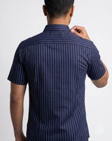 A man viewed from the back wearing a navy blue short-sleeve button-up shirt with thin white vertical pinstripes, set against a plain light gray studio background.