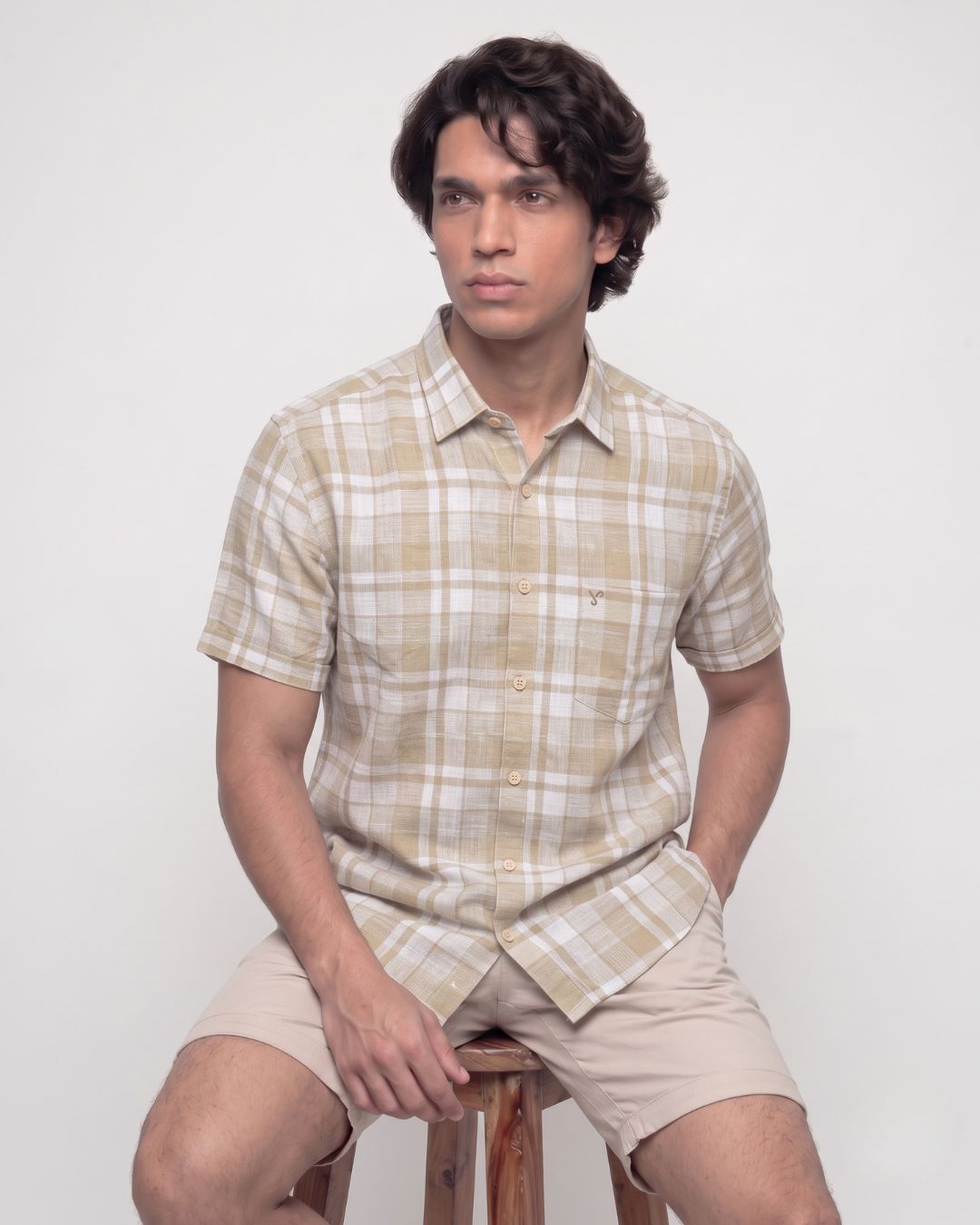 A man wearing a beige and white plaid short-sleeve button-up shirt and matching beige shorts, sitting on a wooden stool against a plain light gray background.