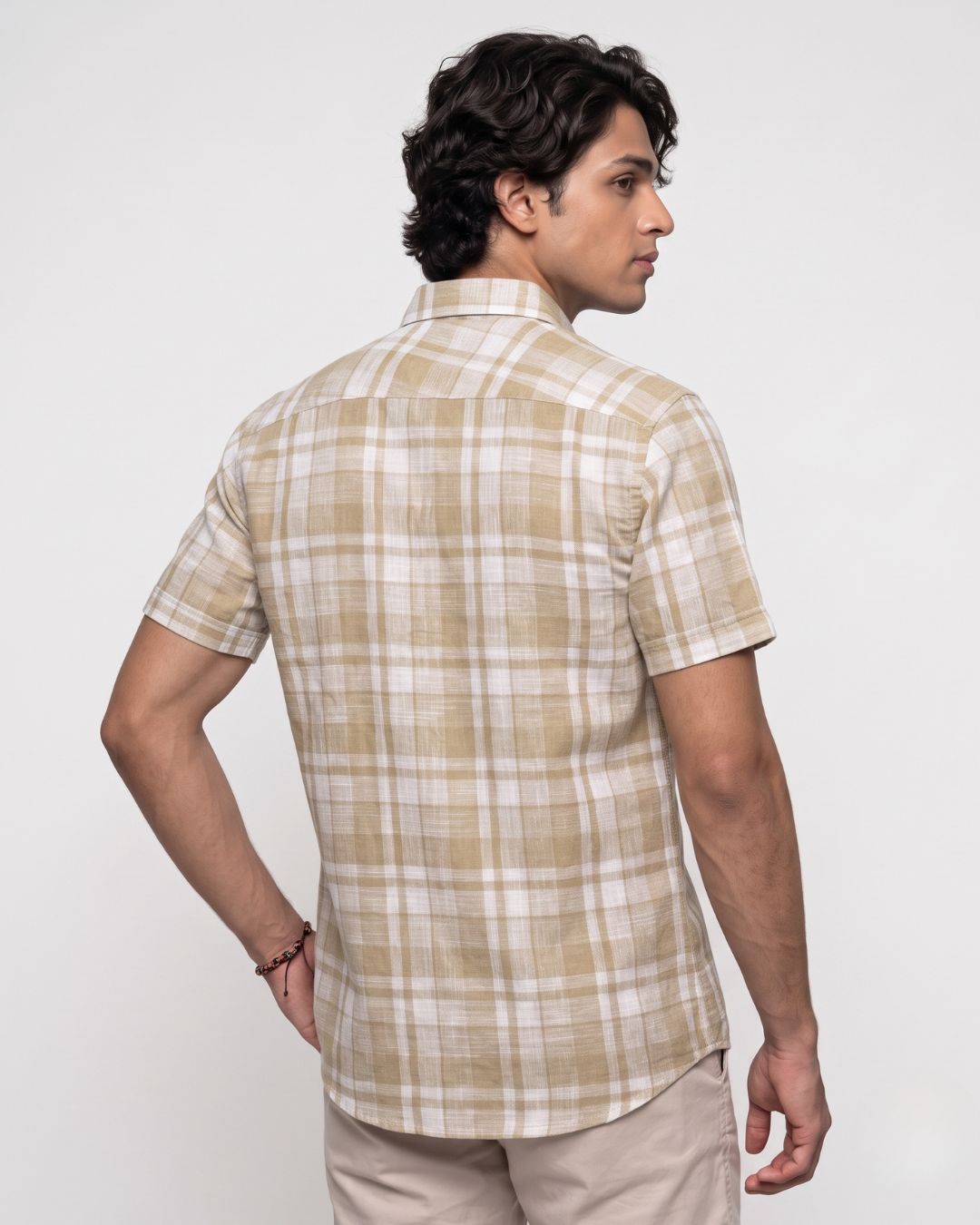 A man wearing a beige and white plaid short-sleeve button-up shirt, shown from a three-quarter back view against a plain light gray studio background.