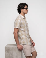 A man in side profile wearing a beige and white plaid short-sleeve button-up shirt and beige shorts, leaning against a concrete block on a light gray background.