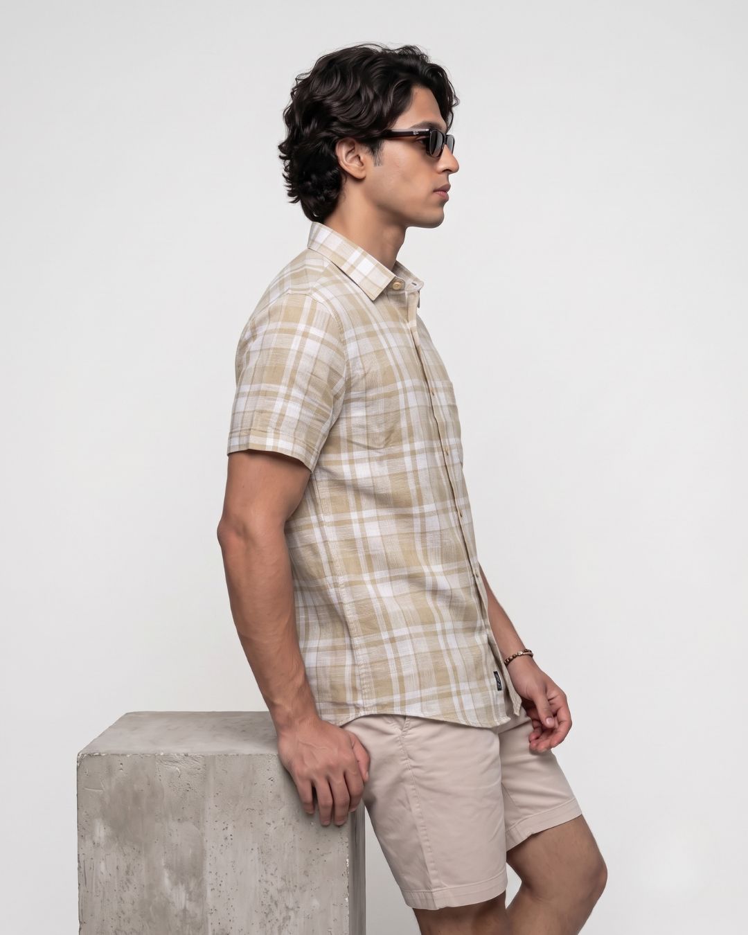 A man in side profile wearing a beige and white plaid short-sleeve button-up shirt and beige shorts, leaning against a concrete block on a light gray background.