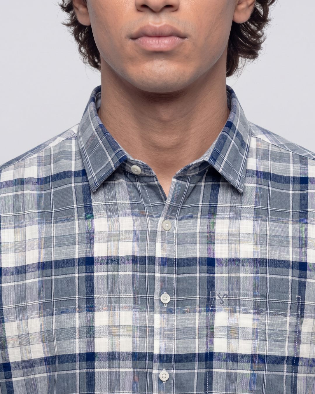 A close-up of a man wearing a blue, white, and gray plaid button-down shirt. The image is cropped from the chin down, showing the collar, buttons, and chest area of the shirt against a plain light gray background.