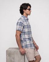 A man in profile wearing a short-sleeve blue and white plaid button-up shirt, beige shorts, and sunglasses, standing next to a concrete block against a light gray studio background.