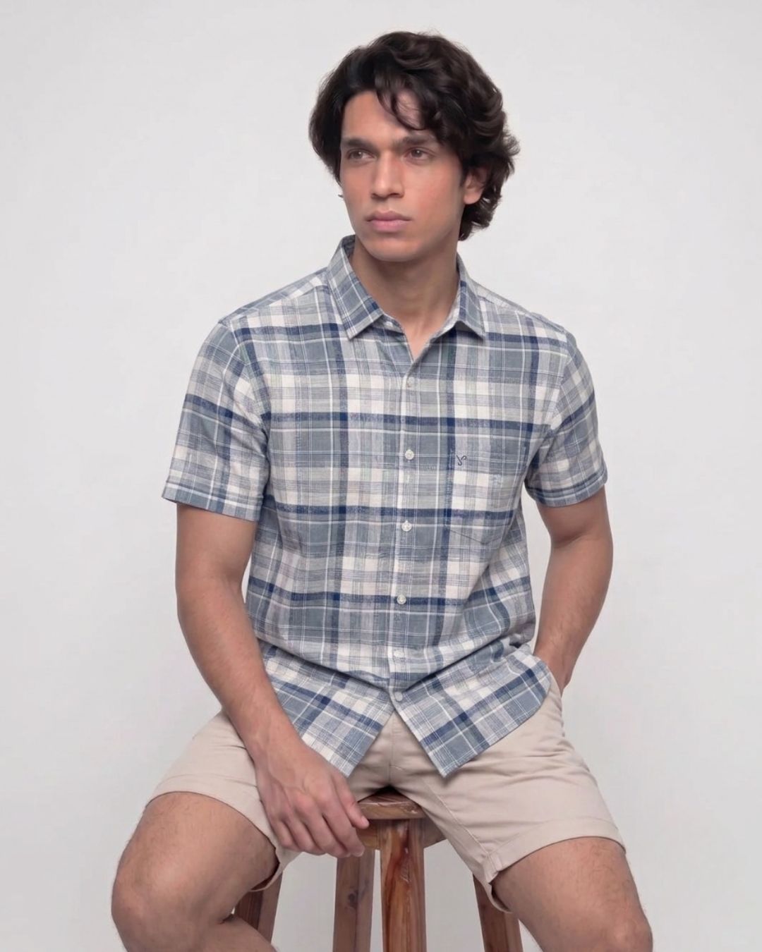 A man wearing a short-sleeve plaid button-up shirt in blue, white, and gray, paired with beige shorts, sitting on a wooden stool against a light gray background.