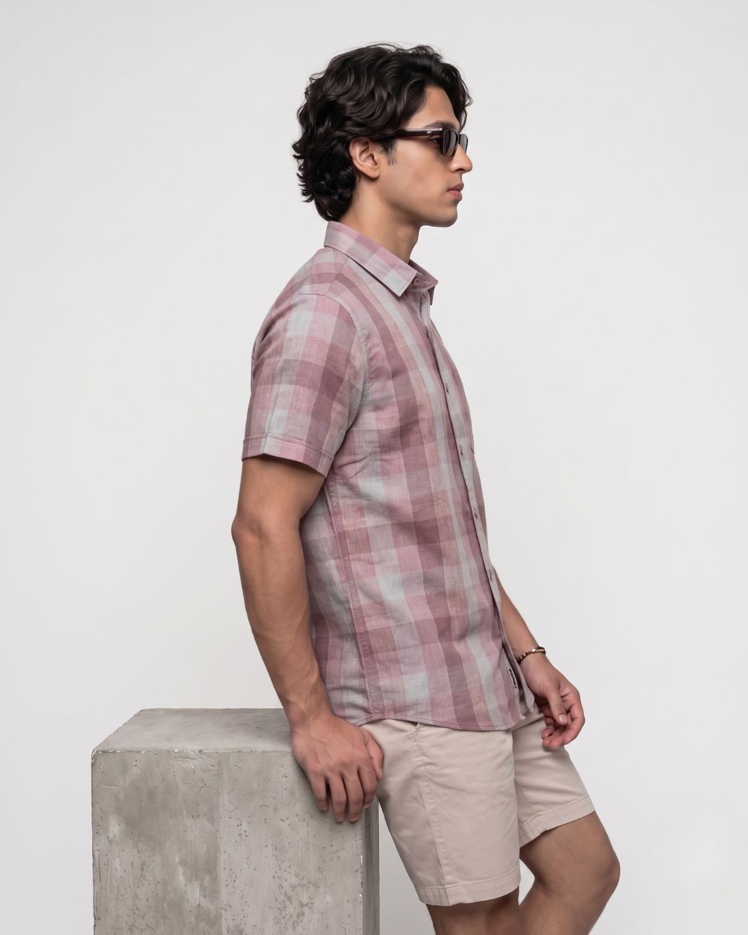 A man in profile wearing a short-sleeve plaid button-up shirt in dusty pink and light gray, paired with beige shorts, standing next to a concrete block against a white studio background.