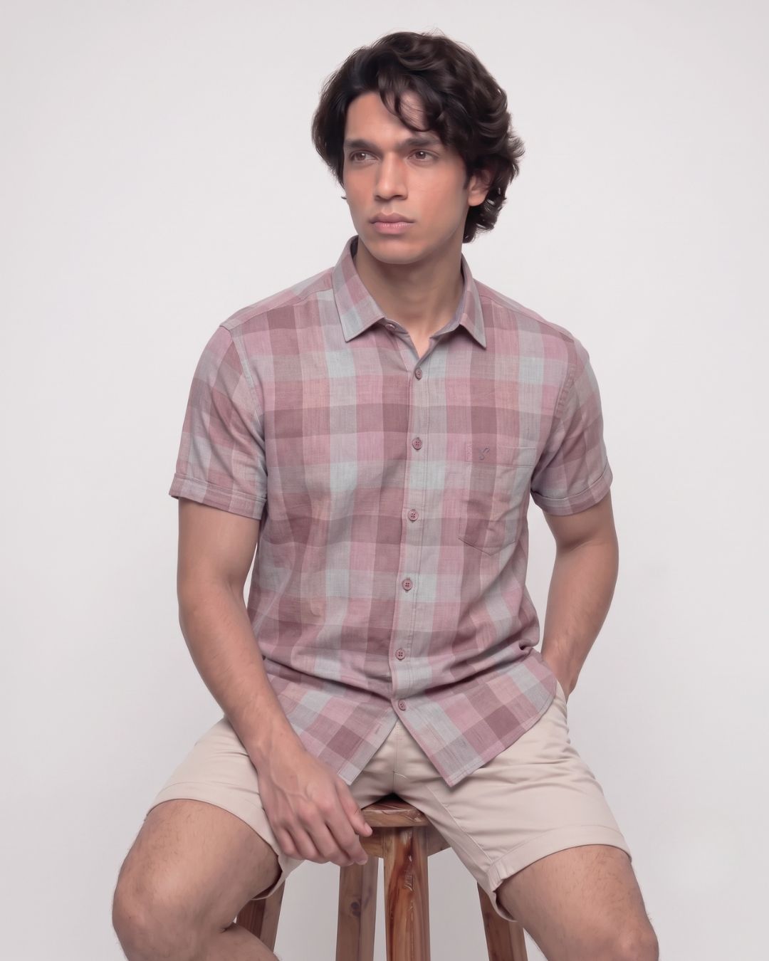 A man wearing a short-sleeve button-up shirt with a dusty pink and light gray plaid pattern, paired with beige shorts, sitting on a wooden stool against a plain white background.