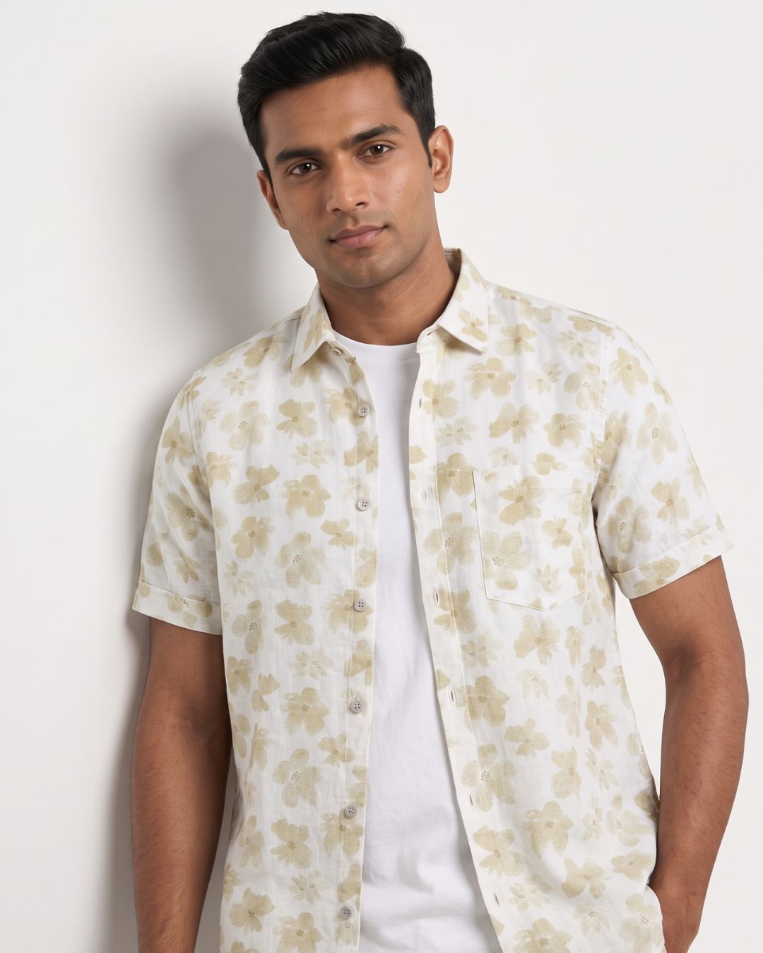 A man wearing a short-sleeve button-up shirt with a beige floral pattern on a white background, layered over a white t-shirt, against a plain off-white wall.