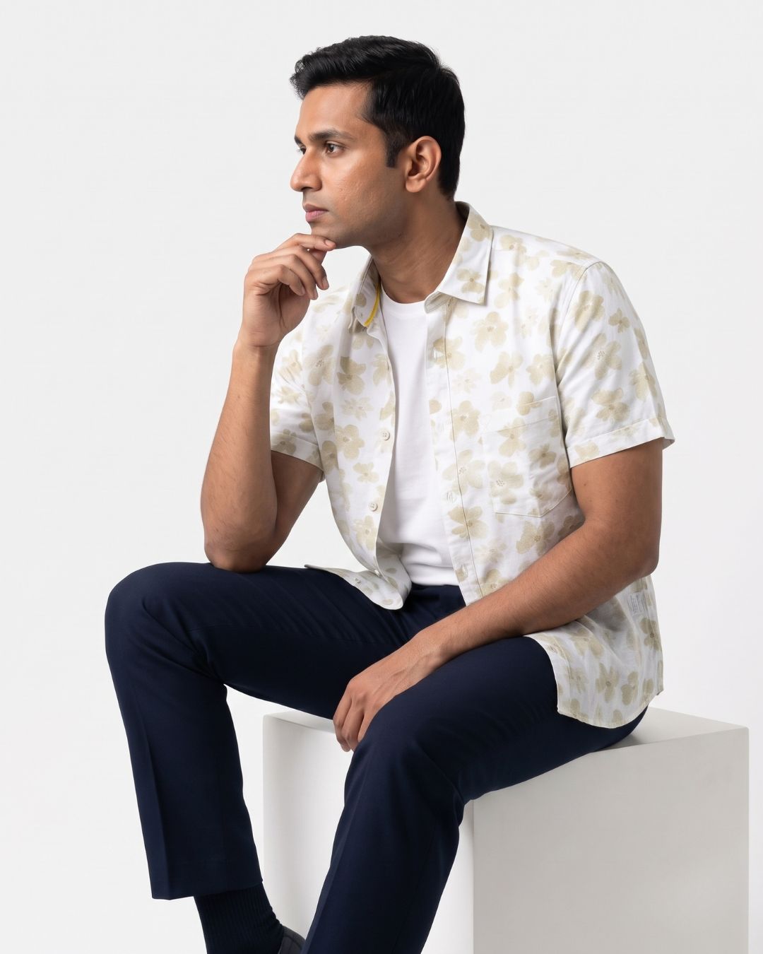 A man sitting on a white block wearing a short-sleeve button-up shirt with a beige and white abstract pattern over a white t-shirt, paired with navy blue trousers, against a plain white background.