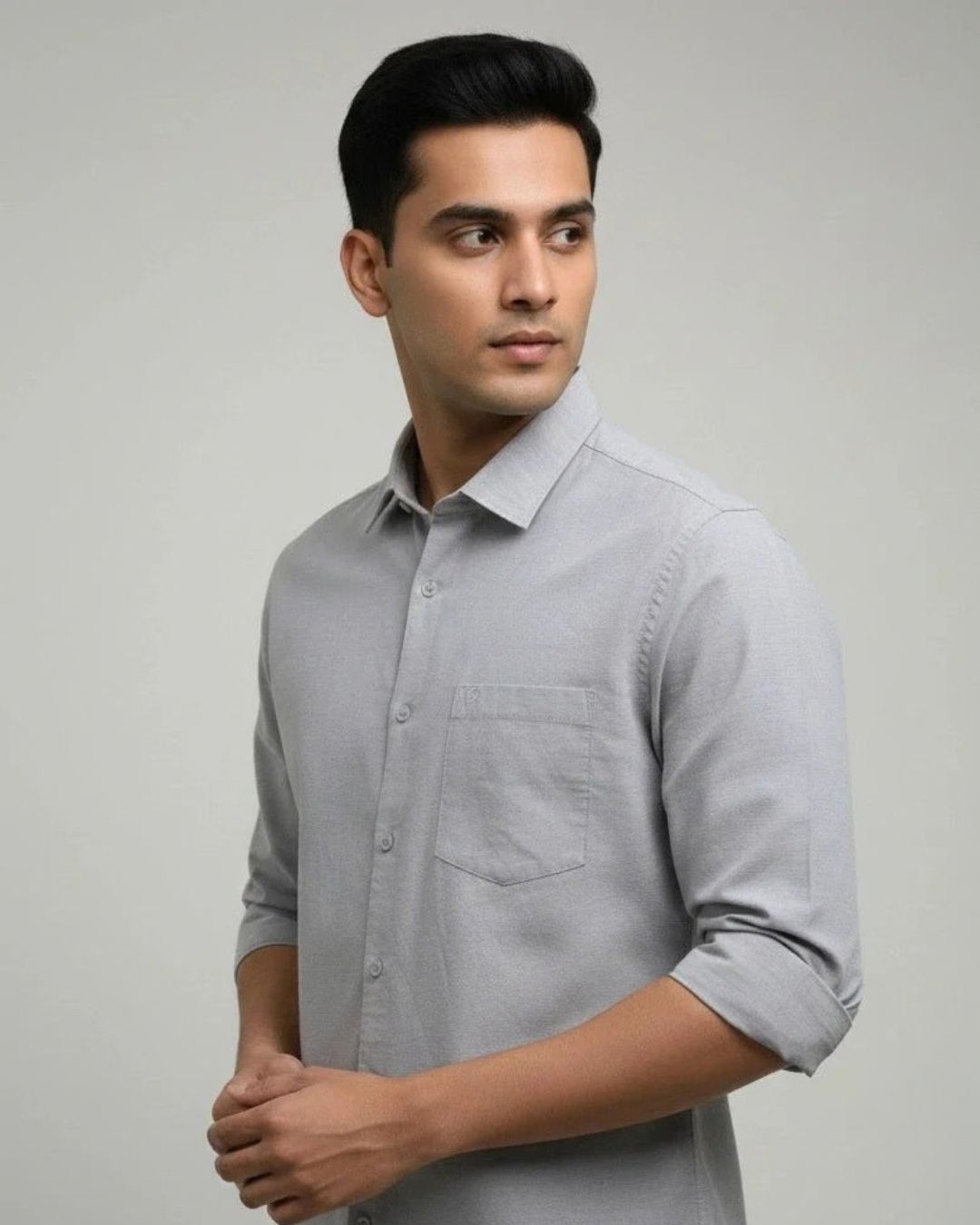 A man wearing a light gray long-sleeve button-down shirt with rolled-up sleeves, standing against a neutral light gray background.
