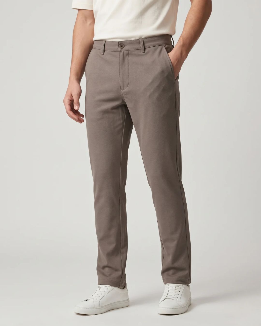 A person wearing slim-fit taupe gray trousers, paired with a cream t-shirt and white sneakers, standing against a light gray studio background.