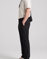 A side-profile view of a person wearing black trousers, a cream short-sleeve polo shirt, and beige sneakers, with a black crossbody bag worn over the shoulder, set against a plain light gray studio background.