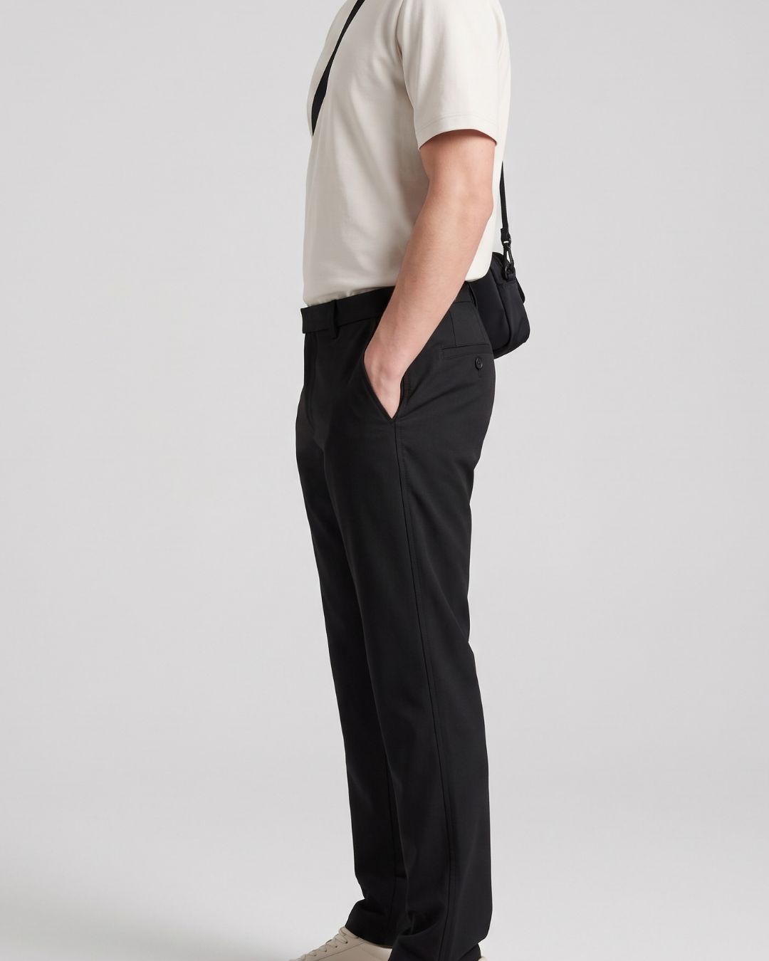 A side-profile view of a person wearing black trousers, a cream short-sleeve polo shirt, and beige sneakers, with a black crossbody bag worn over the shoulder, set against a plain light gray studio background.