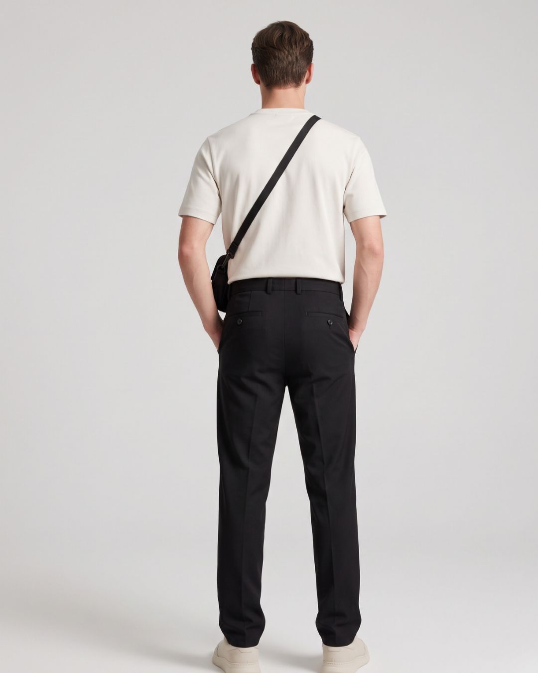 A man viewed from behind wearing a cream short-sleeve t-shirt, black trousers, and cream sneakers, with a black crossbody bag slung over his shoulder, against a light gray studio background.