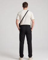 A man viewed from behind wearing a cream short-sleeve t-shirt, black trousers, and cream sneakers, with a black crossbody bag slung over his shoulder, against a light gray studio background.