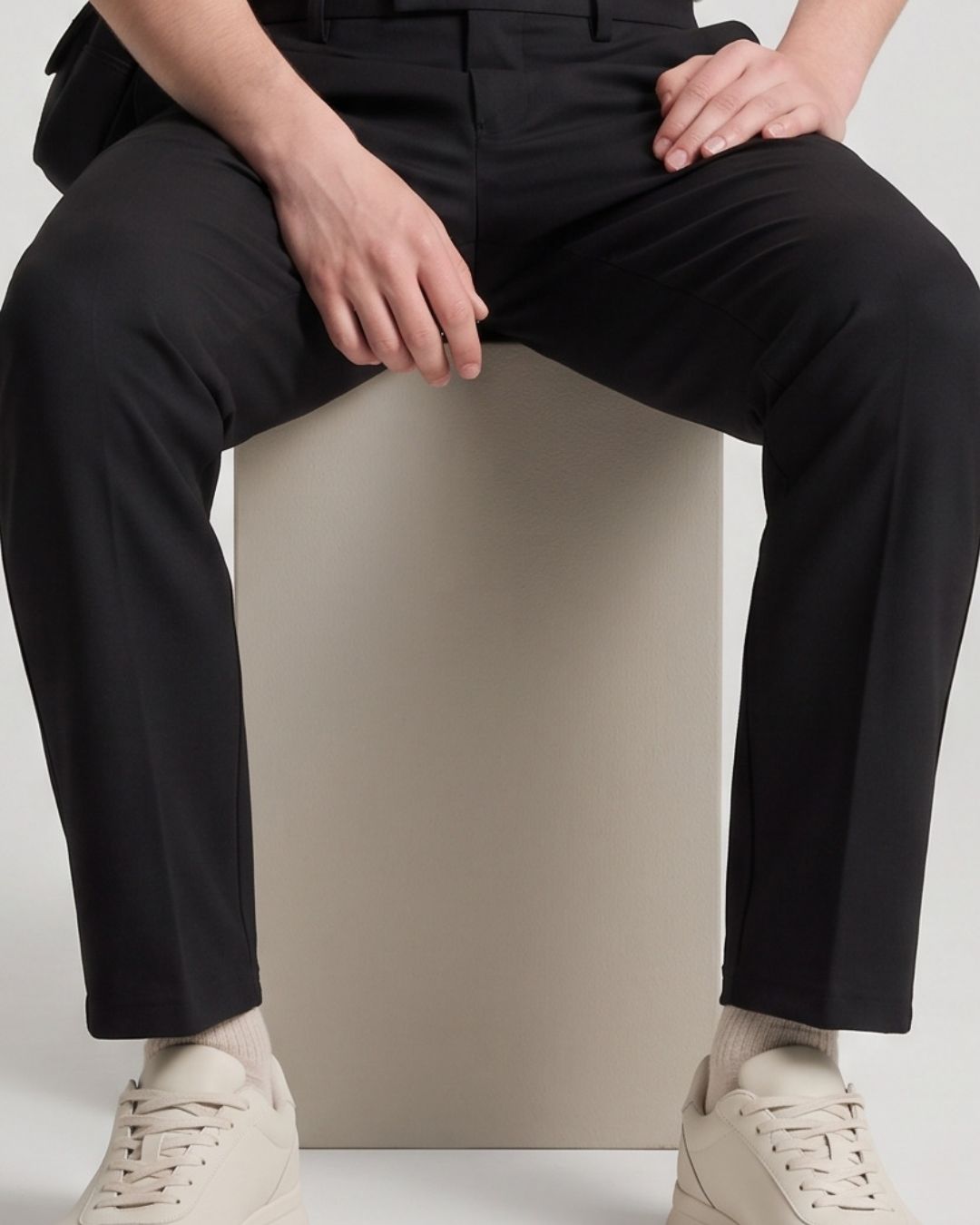A person seated on a beige pedestal wearing slim black trousers, cream socks, and cream sneakers against a light gray studio background.