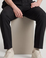 A person seated on a beige pedestal wearing slim black trousers, cream socks, and cream sneakers against a light gray studio background.