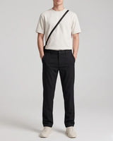 A model wearing a cream short-sleeve crewneck t-shirt, black straight-leg trousers, and off-white sneakers, with a black crossbody bag strap visible across the chest, set against a plain light gray studio background.