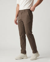 A person wearing slim-fit brown trousers, a white t-shirt, and white sneakers, shown from the waist down against a light gray studio background.
