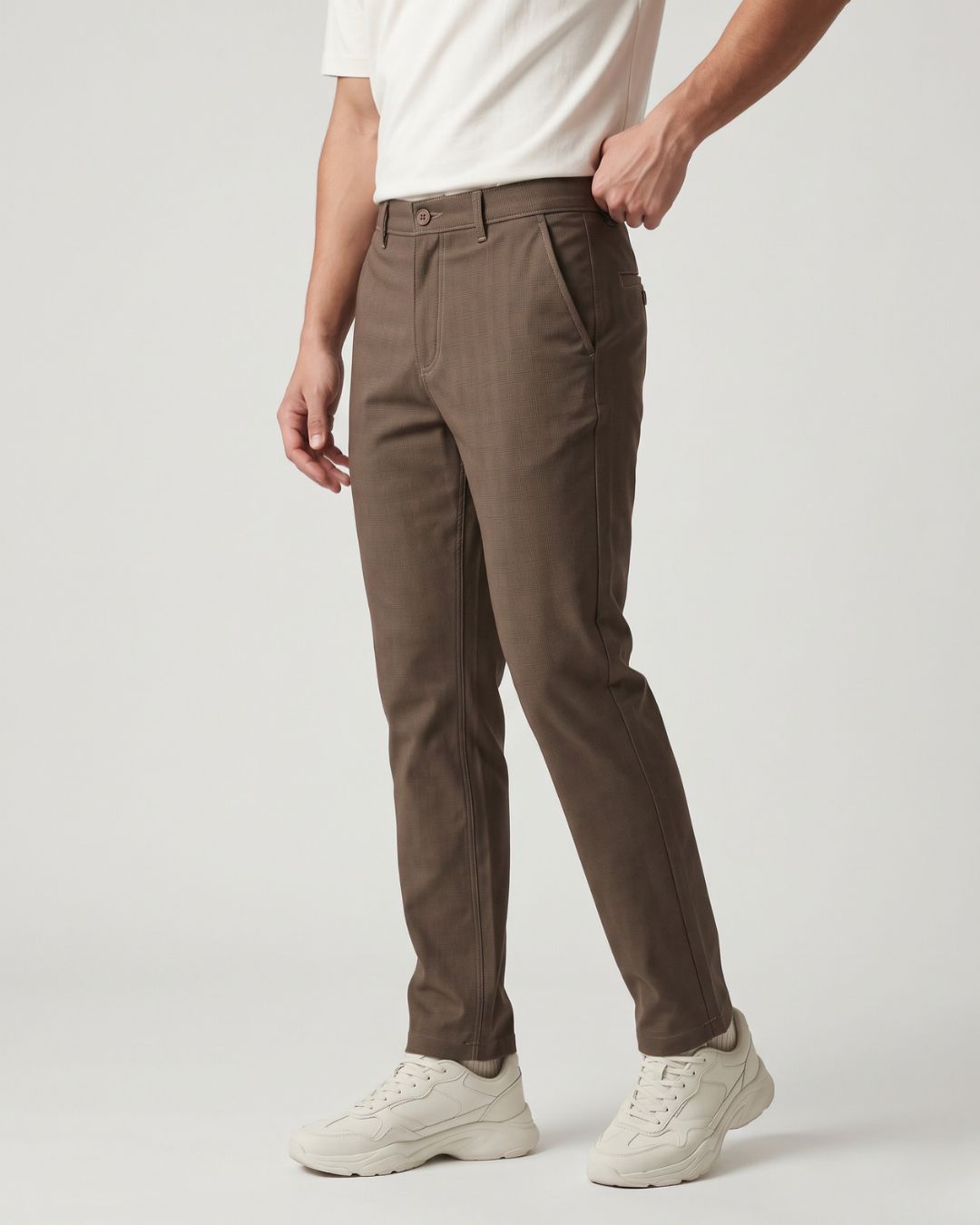 A person wearing slim-fit brown trousers, a white t-shirt, and white sneakers, shown from the waist down against a light gray studio background.