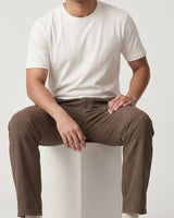 A man seated on a white pedestal wearing a plain white short-sleeve crew-neck T-shirt and brown pants against a light gray studio background.