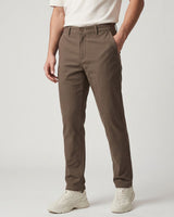 A person wearing slim-fit brown chinos with a white t-shirt and white sneakers, shown from the waist down against a light gray studio background.