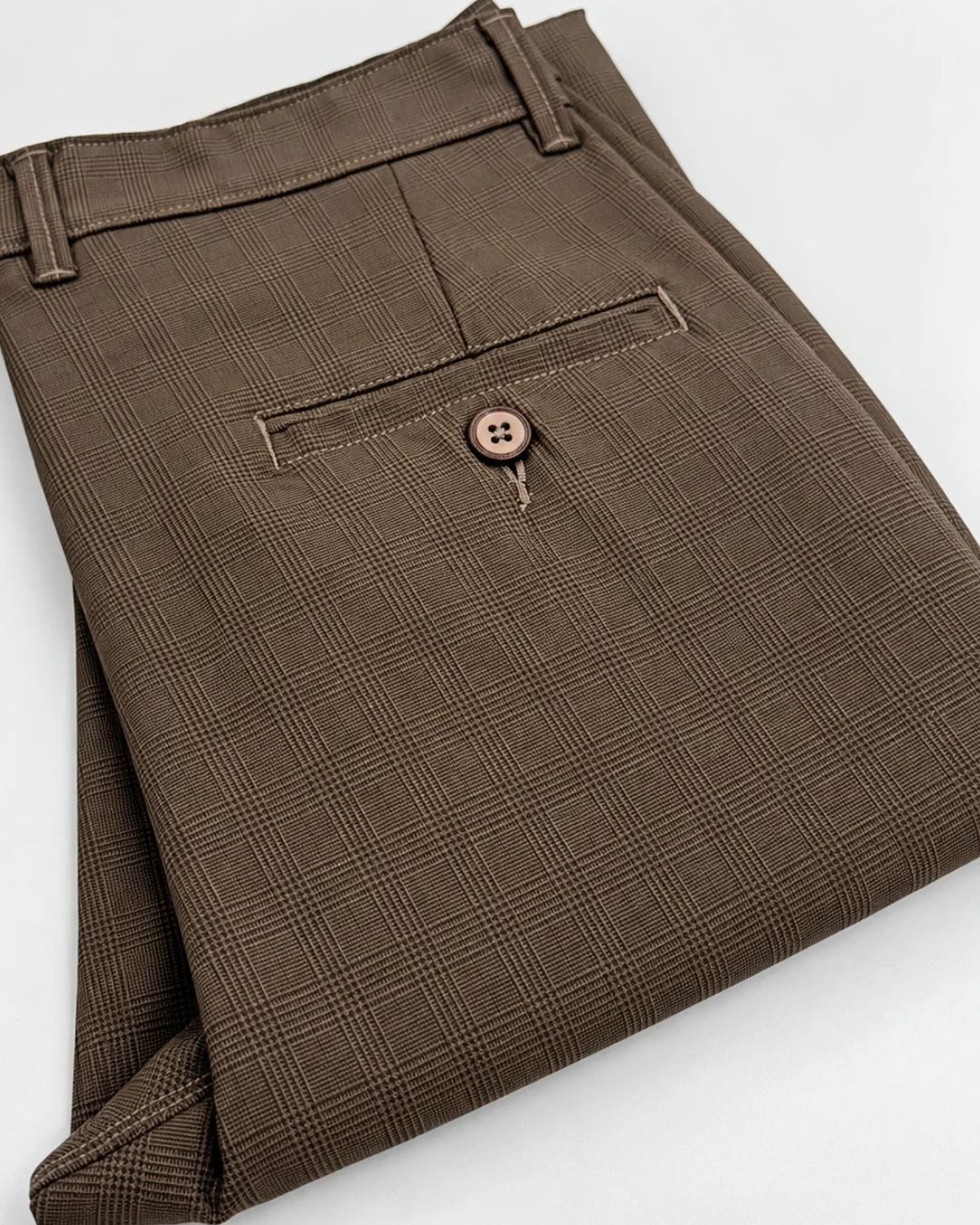 A close-up of folded brown plaid trousers showing the waistband, belt loops, and a rear welt pocket with a decorative button.