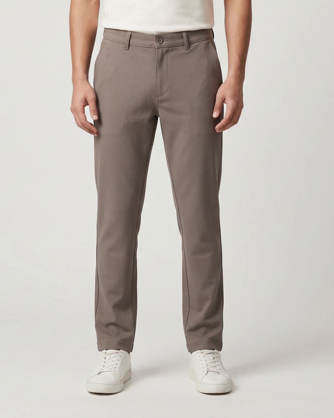 A person wearing slim-fit taupe gray trousers, paired with a white t-shirt and white sneakers, shown from the waist down on a light gray studio background.