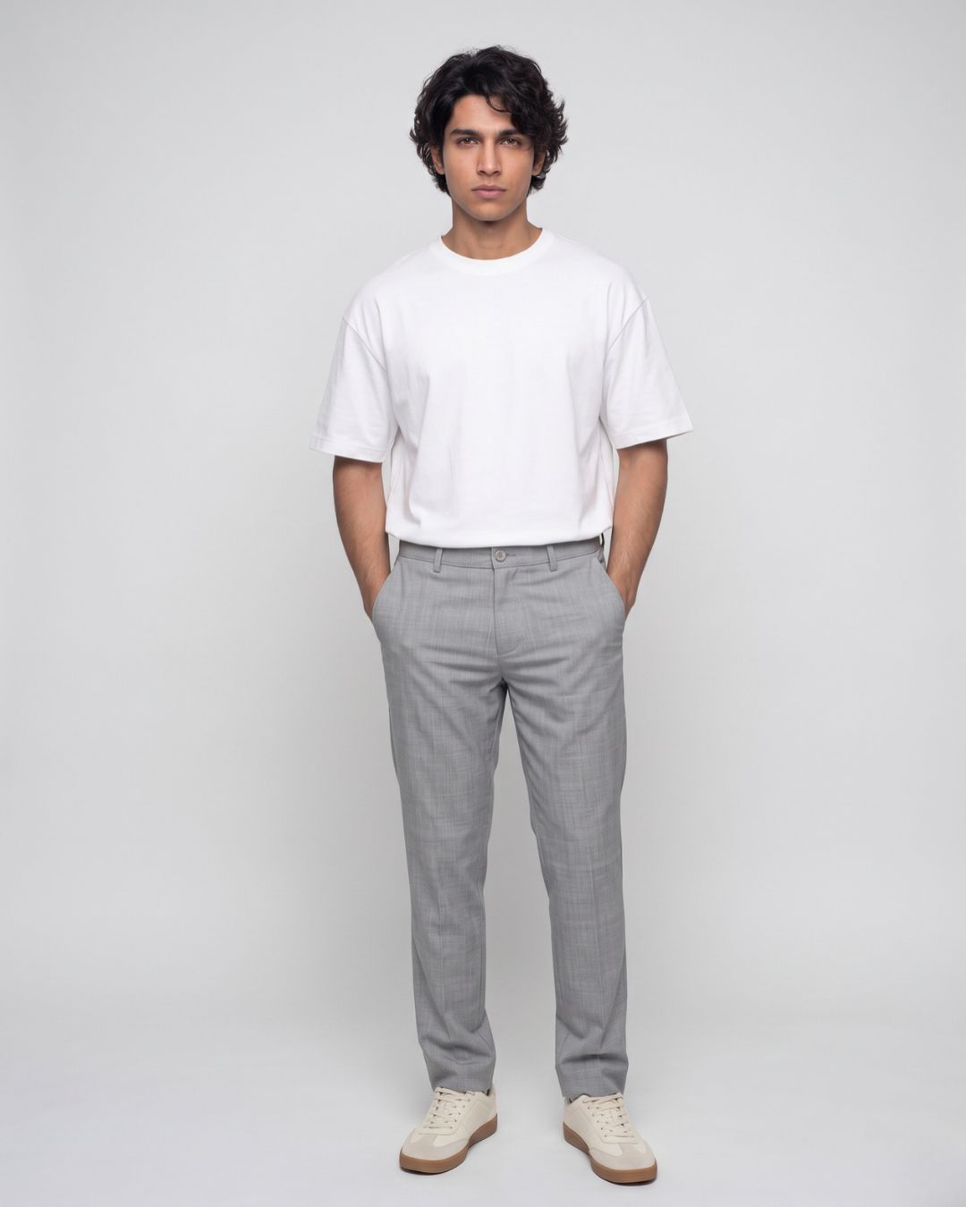 A man standing against a light gray studio background wearing a plain white short-sleeve crewneck t-shirt, light gray trousers, and off-white sneakers.
