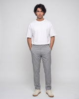 A man standing against a light gray studio background wearing a plain white short-sleeve crewneck t-shirt, light gray trousers, and off-white sneakers.