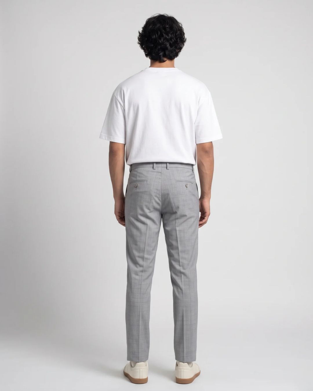 A man standing against a light gray studio background wearing a plain white short-sleeve crewneck t-shirt, light gray trousers, and off-white sneakers.