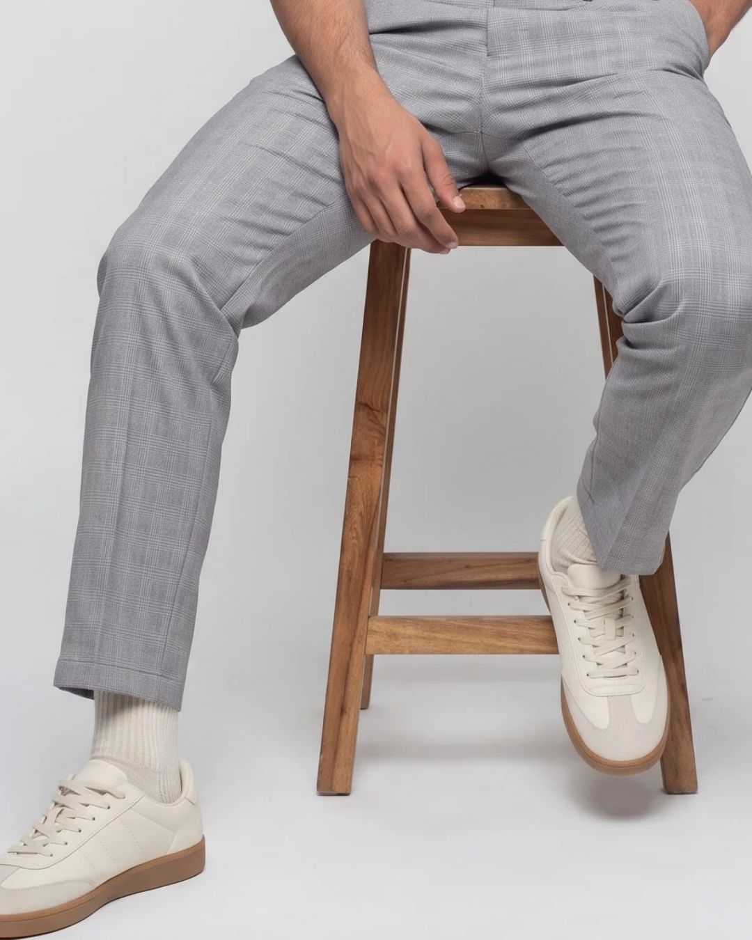 A person seated on a wooden stool wearing light gray plaid trousers, white socks, and off-white sneakers against a plain light gray studio background.