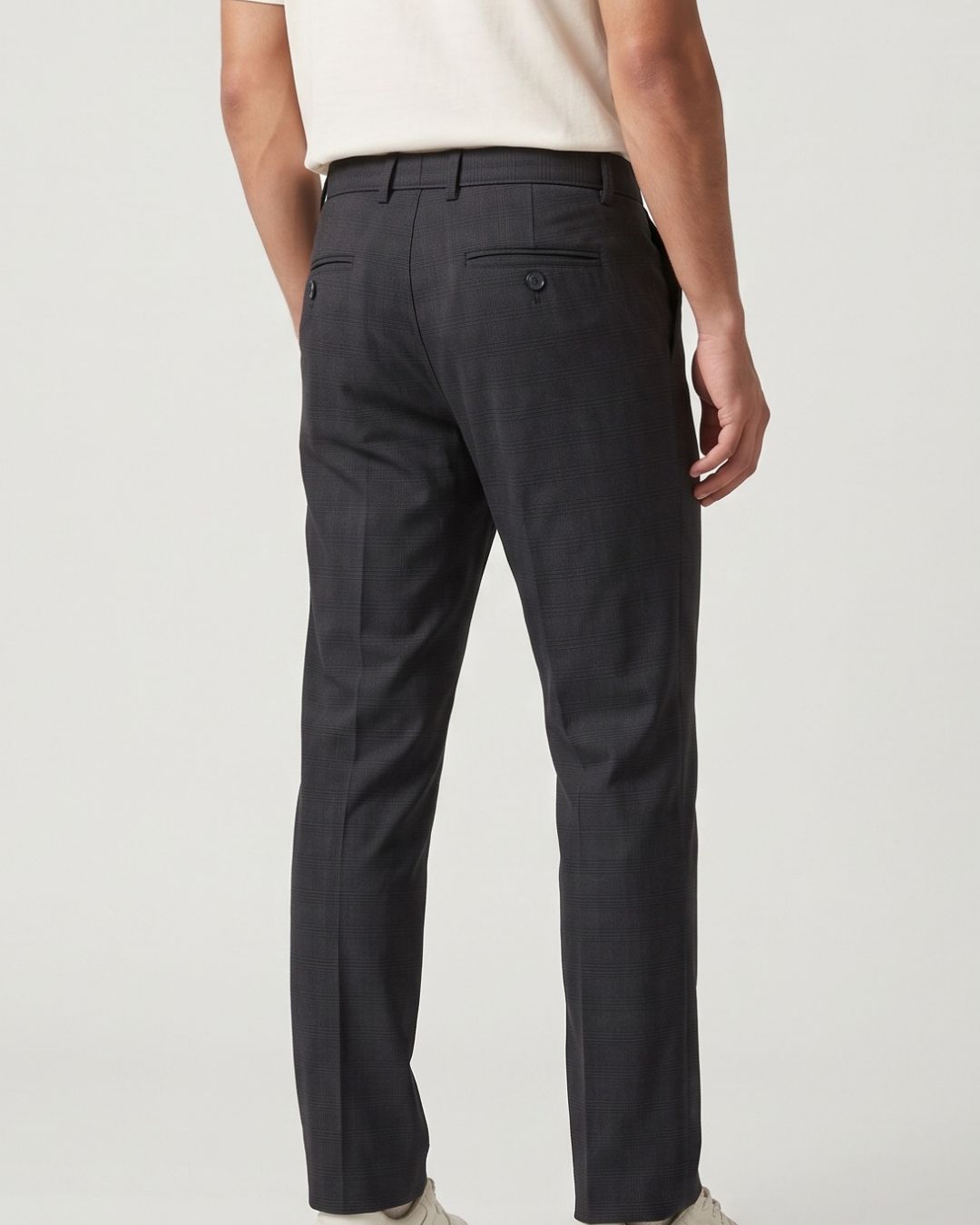 A person shown from the back wearing dark charcoal gray trousers, a cream short-sleeve top, and light-colored shoes against a plain light gray studio background.