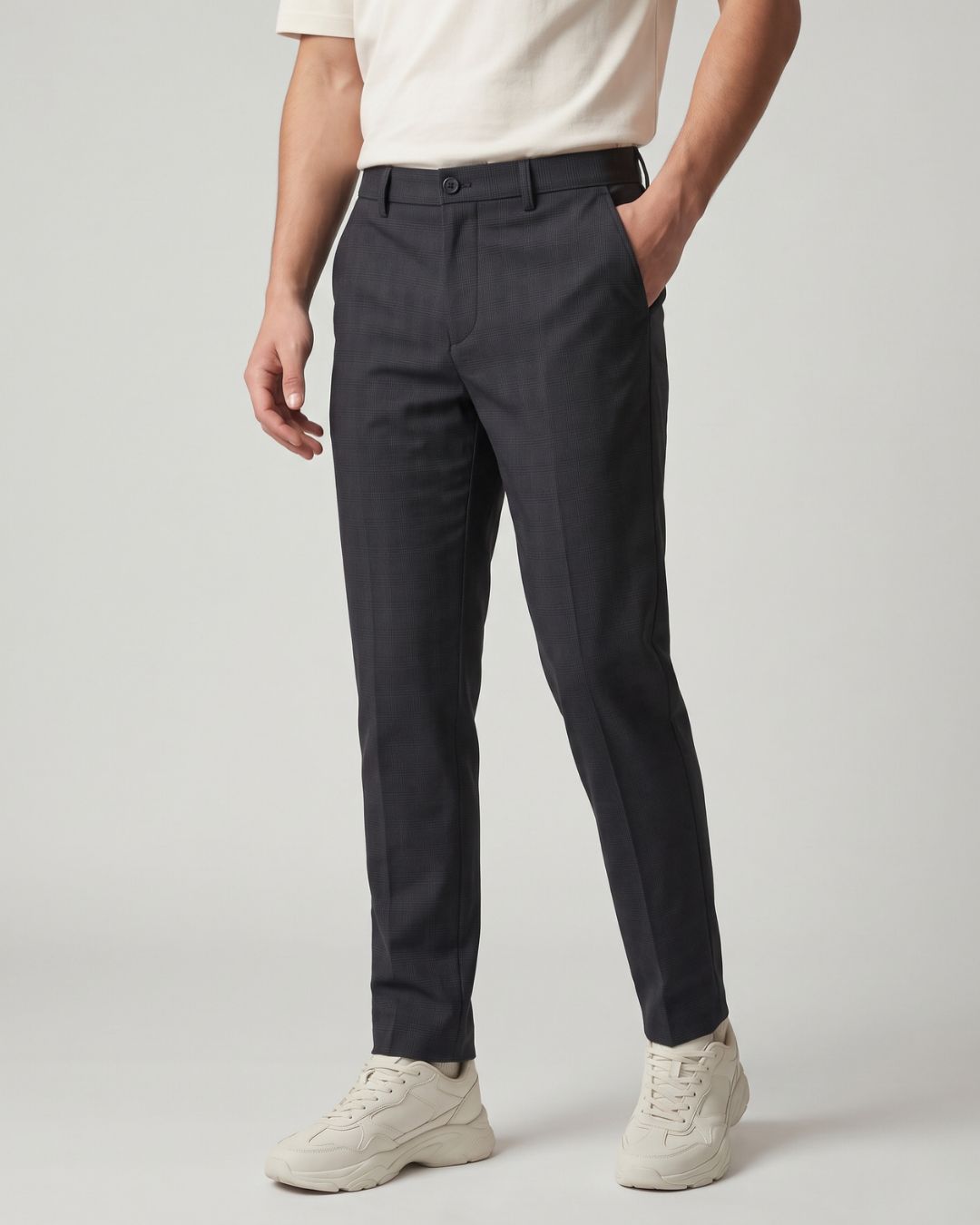 A person wearing dark charcoal gray slim-fit trousers, paired with a cream t-shirt and off-white sneakers, standing against a light gray studio background.