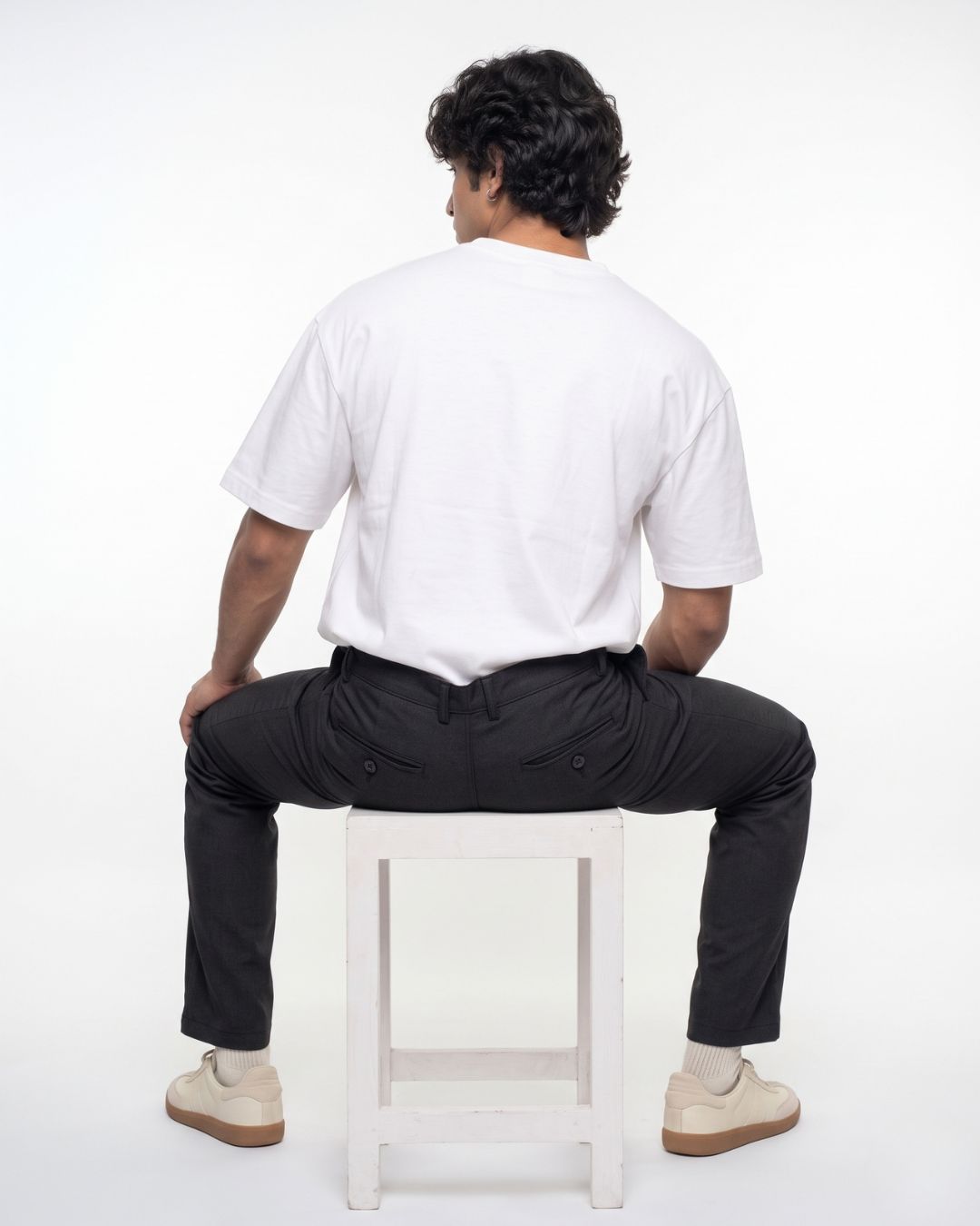 A man is shown from behind, sitting on a white stool. He is wearing a white short-sleeve t-shirt, black trousers, and beige sneakers against a plain white studio background.