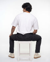 A man is shown from behind, sitting on a white stool. He is wearing a white short-sleeve t-shirt, black trousers, and beige sneakers against a plain white studio background.