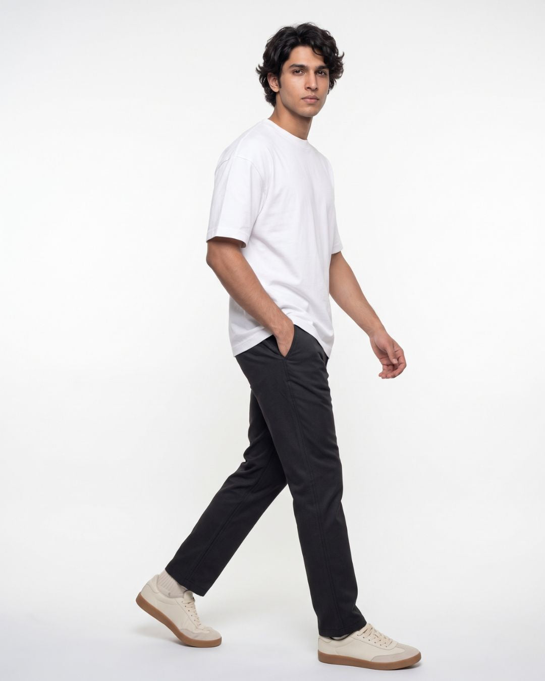 A man wearing a white crew-neck t-shirt, black trousers, and beige sneakers walking in profile against a plain white studio background.