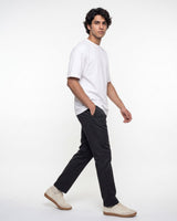 A man wearing a white crew-neck t-shirt, black trousers, and beige sneakers walking in profile against a plain white studio background.