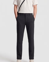 Rear view of a person wearing black tailored trousers with a cream top and a black shoulder bag strap visible against a light gray studio background.