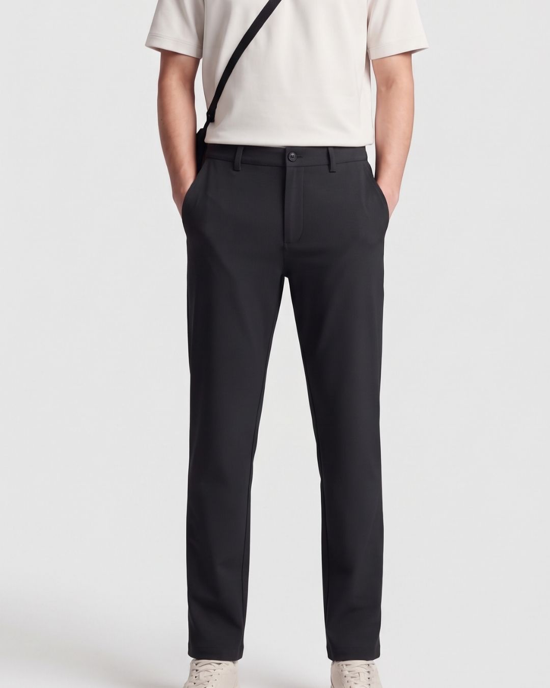 Rear view of a person wearing black tailored trousers with a cream top and a black shoulder bag strap visible against a light gray studio background.