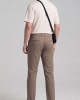 A man shown from behind wearing beige trousers, a cream short-sleeve polo shirt, and carrying a small black crossbody pouch against a light gray studio background.