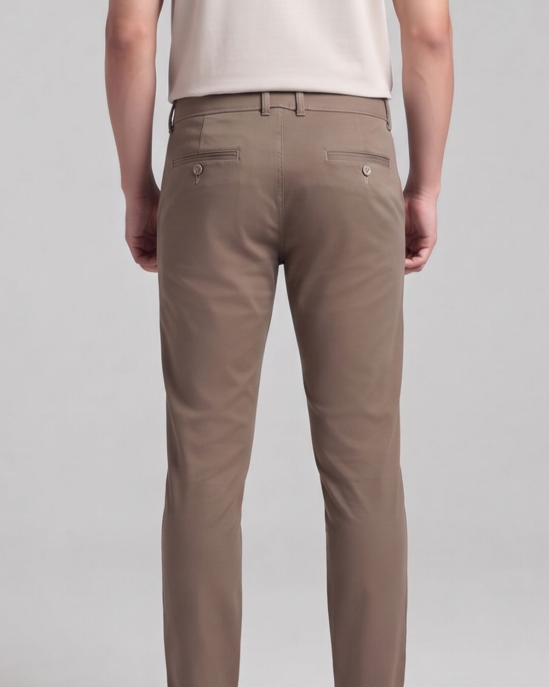 A person shown from the back wearing slim-fit trousers in a warm taupe-brown color, paired with a light cream top, against a neutral gray studio background.