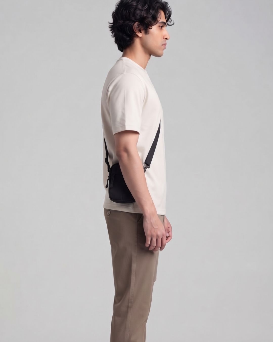 A man in a side profile wearing a cream t-shirt, beige trousers, and a black crossbody bag with a thin black strap, set against a plain light gray studio background.