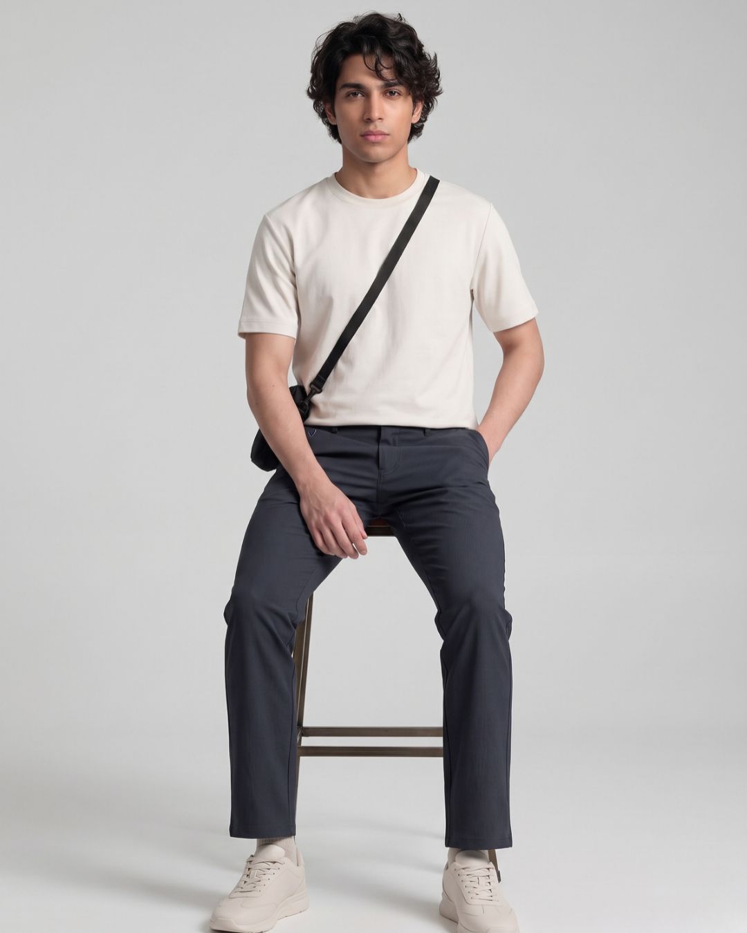 A man sitting on a stool wearing a cream crew-neck t-shirt, dark gray trousers, and off-white sneakers, with a black crossbody bag slung over his shoulder, against a light gray studio background.