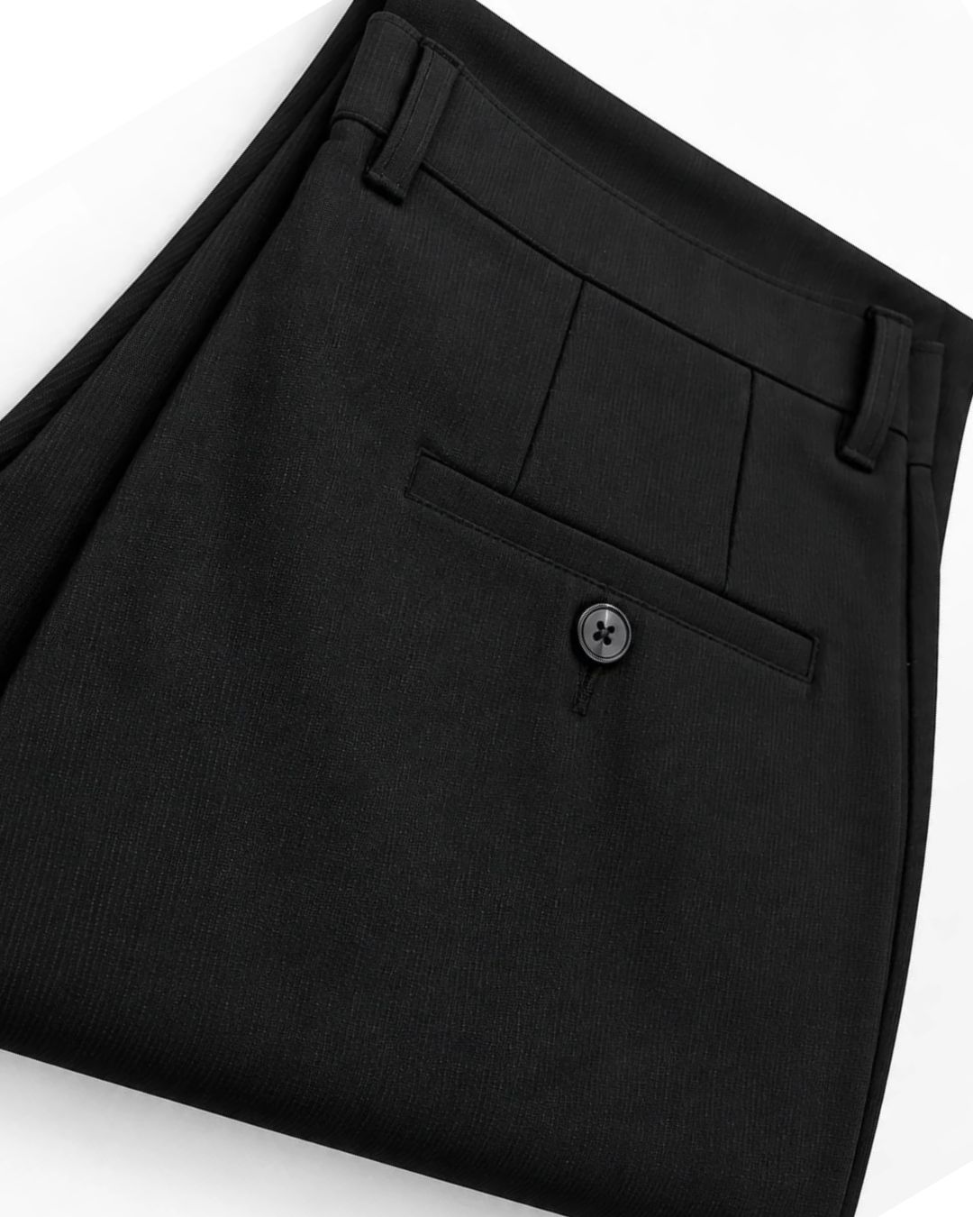 Close-up of folded black trousers showing the waistband, belt loops, front flap pocket with a button, and fabric texture on a white background.
