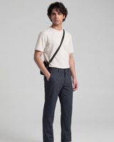 A man modeling a cream short-sleeve crewneck t-shirt, dark gray trousers, and a black crossbody bag with a thin black strap, standing against a light gray studio background.