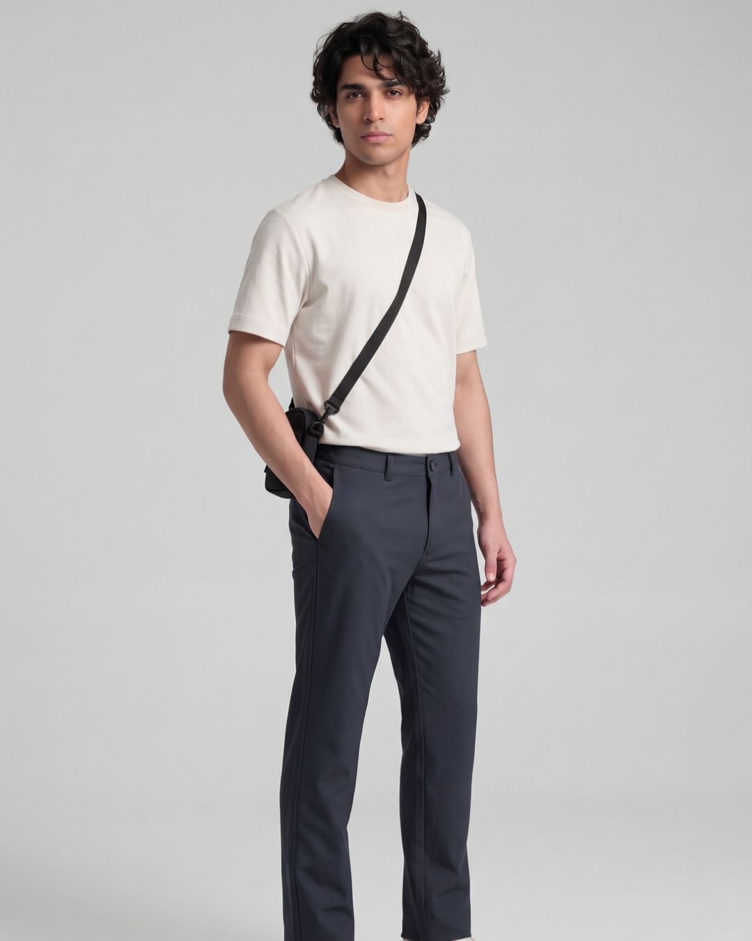 A man modeling a cream short-sleeve crewneck t-shirt, dark gray trousers, and a black crossbody bag with a thin black strap, standing against a light gray studio background.