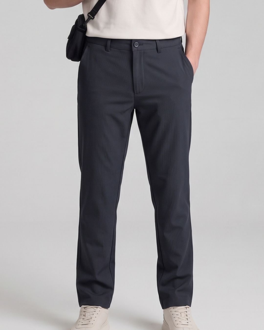 A person wearing dark navy straight-leg dress pants, styled with a cream t-shirt, a black crossbody bag, and off-white sneakers against a light gray studio background.