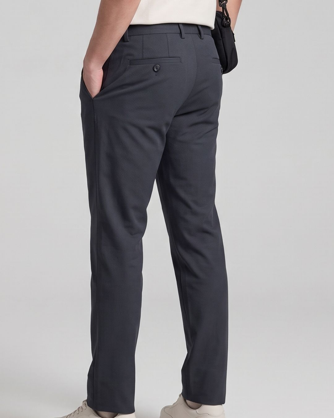 A person shown from the back wearing dark charcoal gray slim-fit trousers, a white top, and light beige sneakers, with a small black shoulder bag hanging at the side, against a light gray studio background.