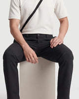 Rear view of a person wearing black tailored trousers with a cream top and a black shoulder bag strap visible against a light gray studio background.