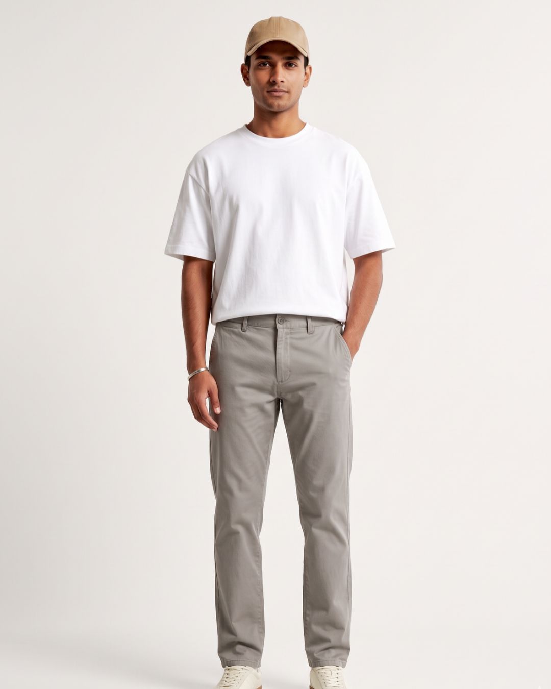 A man modeling a white crewneck t-shirt and light gray chino pants, paired with a beige baseball cap and white sneakers, against a plain off-white studio background.